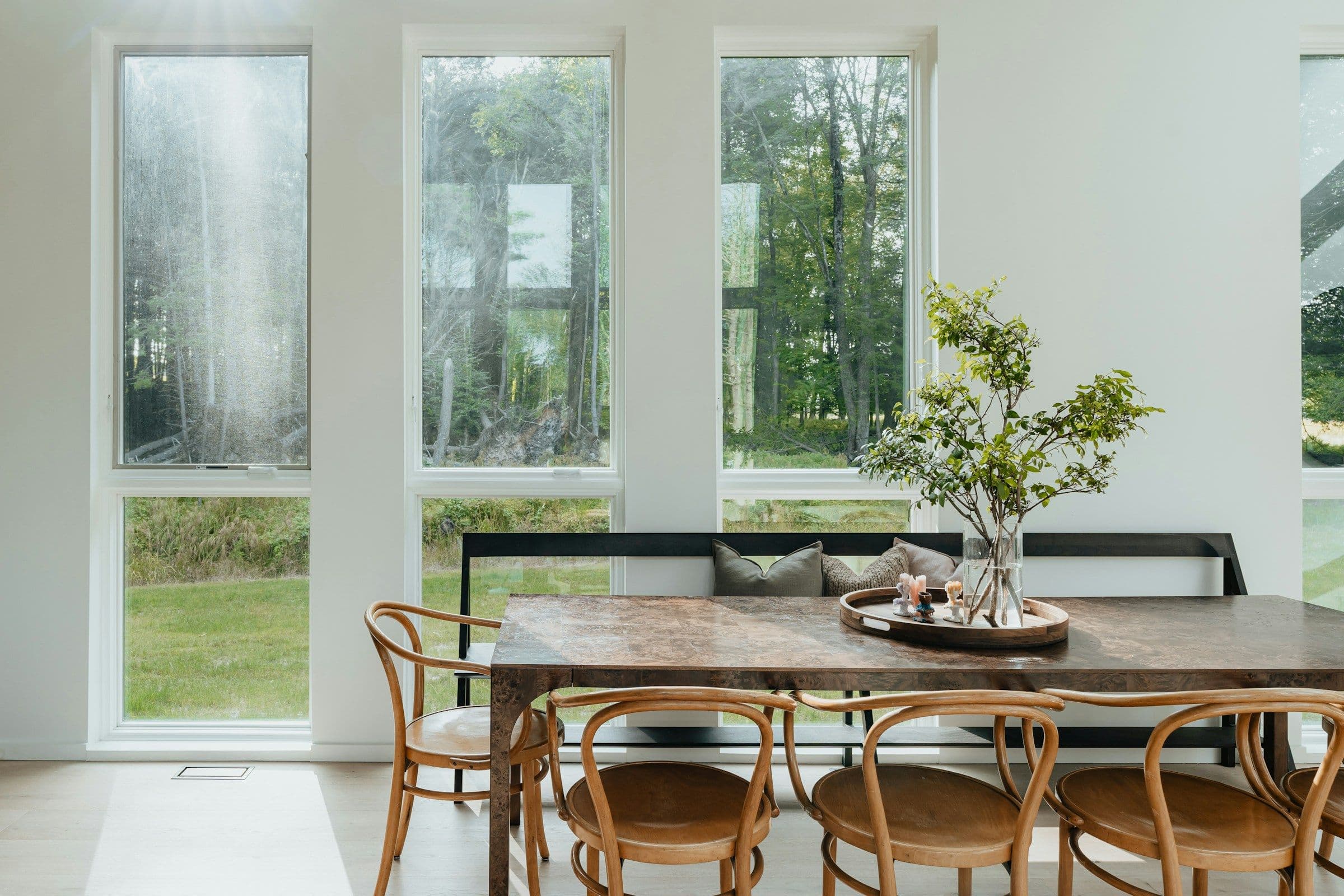 Salle à manger lumineuse : grandes fenêtres sur la verdure, table en pierre, chaises bistrot en bois courbé et branchages dans un vase.