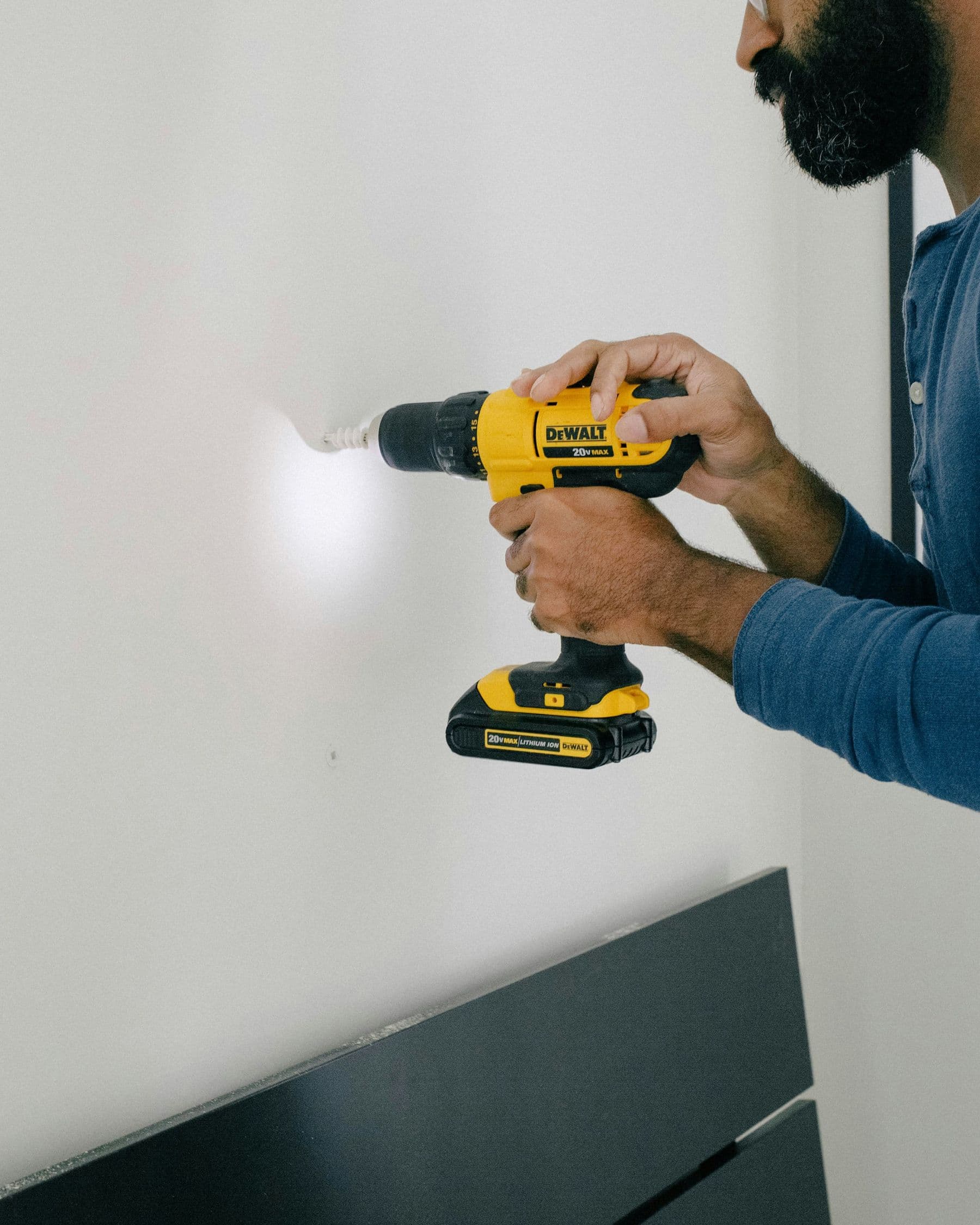Un homme perce un mur blanc avec une perceuse-visseuse jaune, pour y poser une cheville.