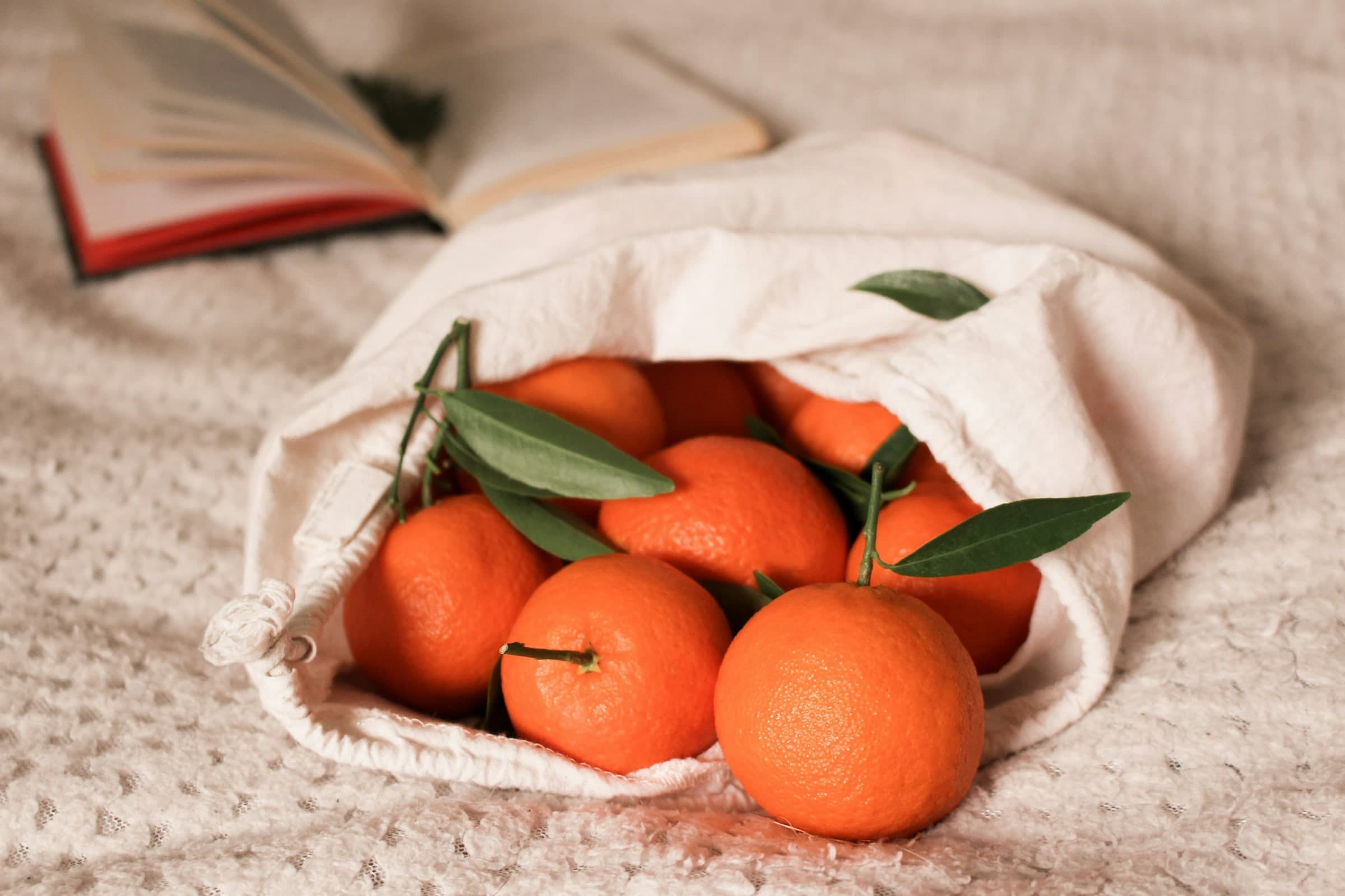Un sac en coton écru rempli d’oranges feuillées, posé sur un plaid clair.