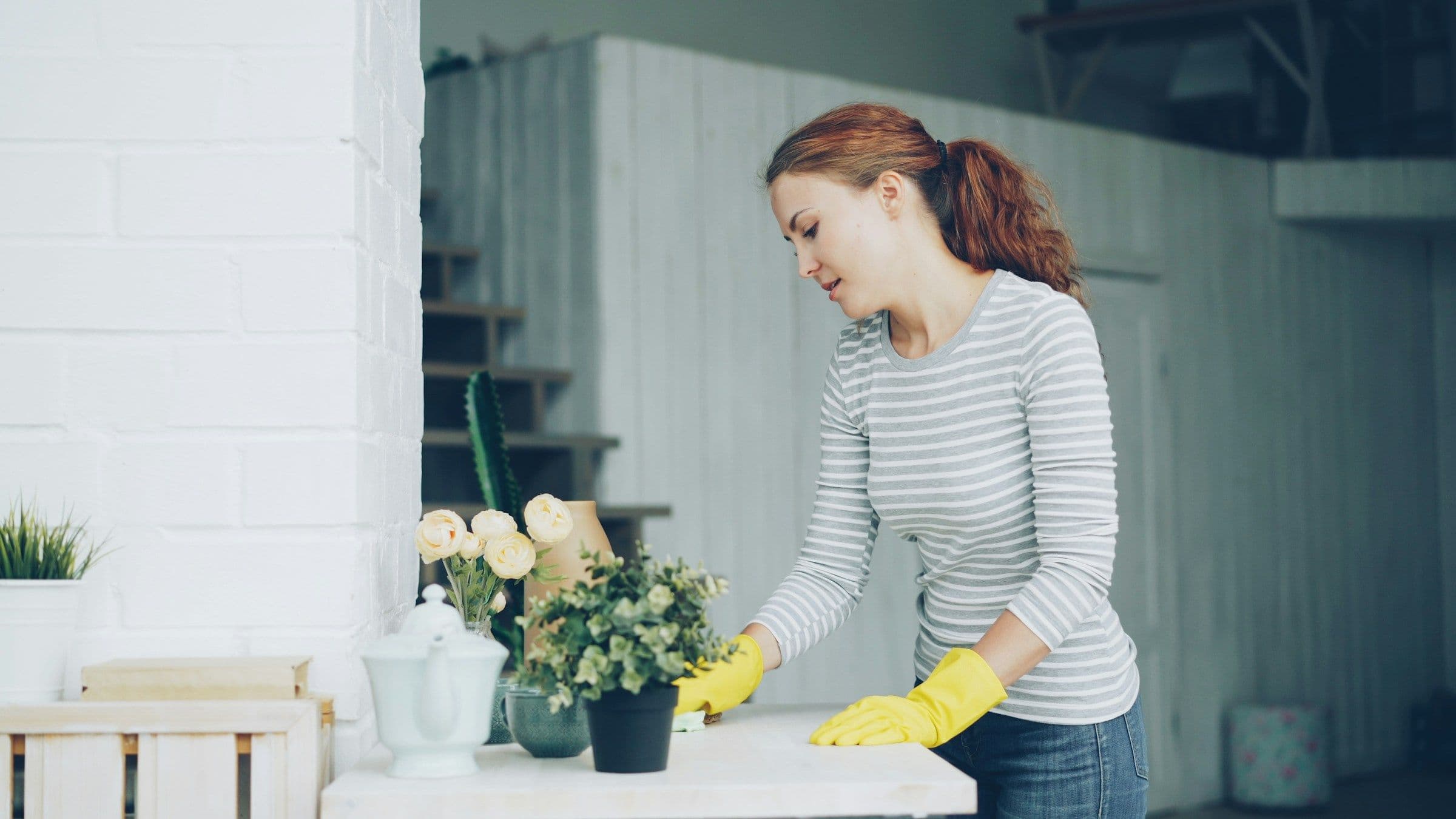 Une femme en gants jaunes essuie une table blanche près d’un bouquet, dans un intérieur clair.