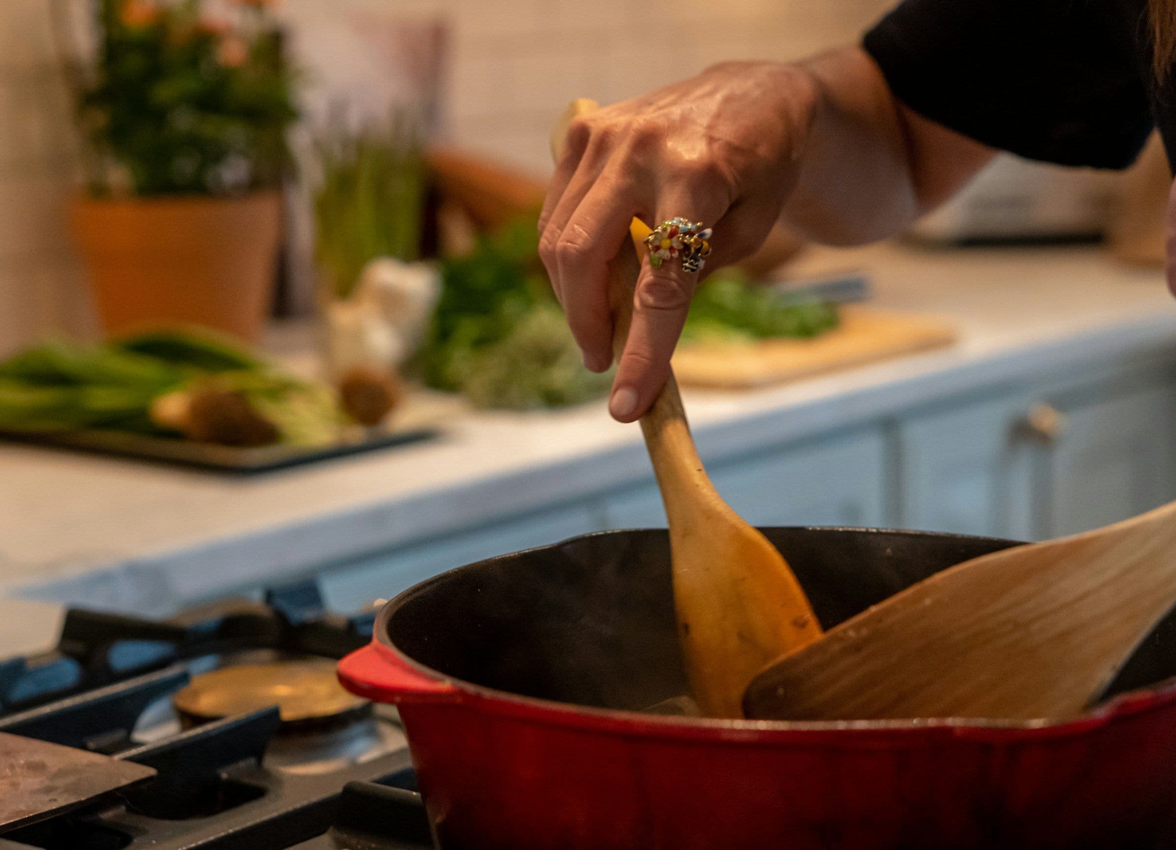 Une main remue à la cuillère en bois le contenu d’une cocotte rouge, devant un plan de travail garni de légumes.